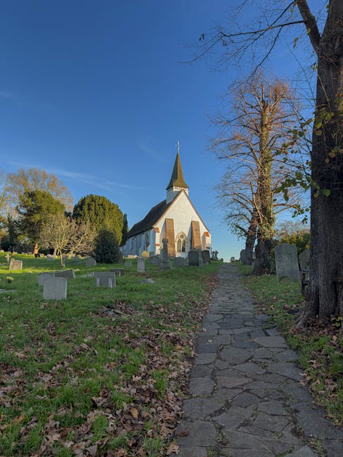 A narrow, uneven stone pathway leading uphill through a cemetery with grass and fallen leaves on either side, alongside a large, leafless tree on the right. In the background, a white church with a tall, pointed steeple is visible under a clear blue sky. The scene is illuminated by natural daylight, suggesting a calm, bright day. This setting could be part of a home relocation or moving logistics context, where careful handling of belongings is necessary before or after transport by Man with Van West Drayton, as seen on their website page about removals quotes and fees.