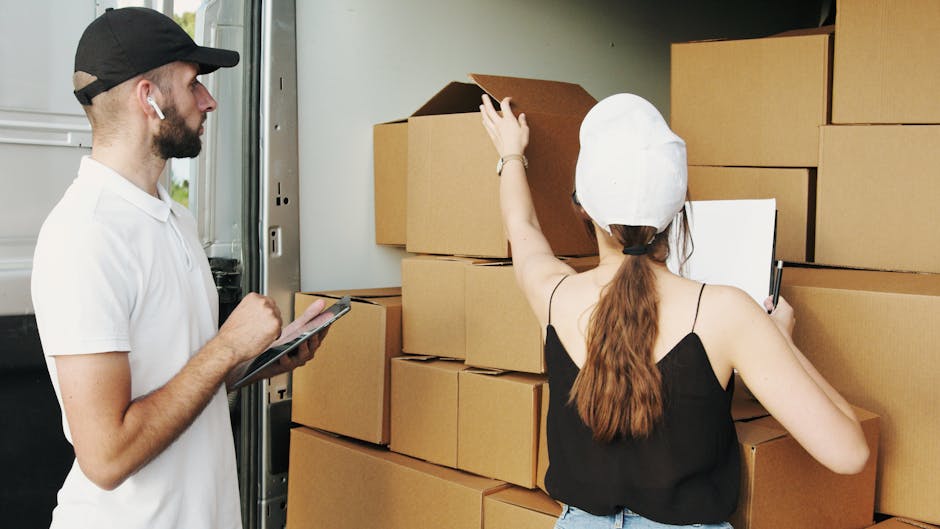 A man and a woman are engaged in a home relocation process inside a moving van. The man, dressed in a white polo shirt and black cap, is standing beside the open vehicle door, holding a clipboard and pen, likely overseeing the packing or loading of boxes. The woman, wearing a white cap, black sleeveless top, and jeans, is positioned with her back to the camera, reaching up to adjust or inspect stacked cardboard boxes of various sizes inside the van. These boxes are arranged on the vehicle’s floor and a small ledge, some open with flaps folded back, indicating active packing or inventory checking. The scene is illuminated by natural light entering through the van's open side, with additional packing materials such as plastic wrap or packing paper possibly inside the boxes not visible. This image exemplifies the loading process typically involved in furniture transport, packing and moving services, and house removals, as provided by Man with Van West Drayton, supporting efficient and organized home relocation.