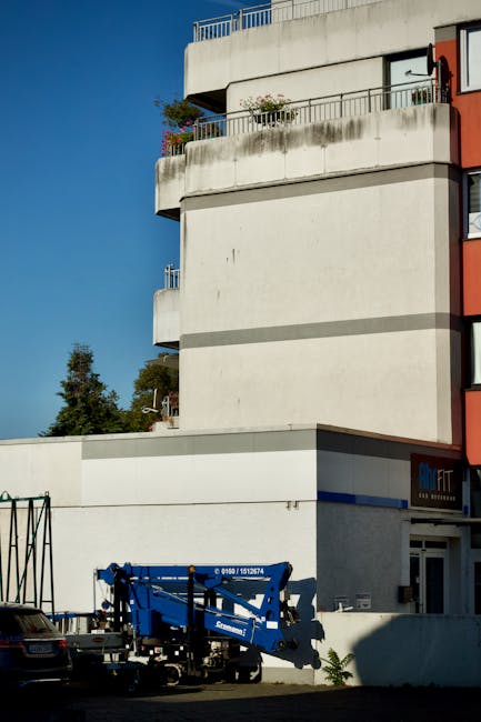 A multi-storey residential building with white exterior walls and rounded balconies featuring metal railings, situated in West Drayton. A blue mobile crane or lift platform, branded 'Genie,' is positioned at ground level near the building, likely used for home relocation or furniture transport. The lifting equipment is on a trailer with stabilising jacks, and nearby vehicles are parked on the street. Bright daylight and clear blue sky illuminate the scene, with some greenery visible in the background. This setup suggests ongoing moving or packing and moving activities, possibly related to house removals facilitated by Man with Van West Drayton. The environment shows an urban setting with a focus on the logistics of indoor furniture and box transportation to upper floors.