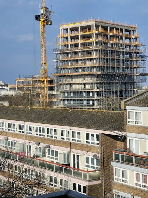 A multi-storey residential building with white exterior walls and rounded balconies featuring metal railings, situated in West Drayton. A blue mobile crane or lift platform, branded 'Genie,' is positioned at ground level near the building, likely used for home relocation or furniture transport. The lifting equipment is on a trailer with stabilising jacks, and nearby vehicles are parked on the street. Bright daylight and clear blue sky illuminate the scene, with some greenery visible in the background. This setup suggests ongoing moving or packing and moving activities, possibly related to house removals facilitated by Man with Van West Drayton. The environment shows an urban setting with a focus on the logistics of indoor furniture and box transportation to upper floors.