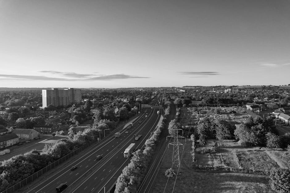 A black and white aerial photograph showing a busy suburban road with multiple lanes of moving vehicles, including cars, a bus, and trucks. The row of trees and residential houses line both sides of the road, with some houses partially obscured by foliage. To the right of the road, there are open grassy fields and small, fenced-off plots, with a few buildings scattered in the distance. The sky above is mostly clear with a few scattered clouds. In the foreground, a high-voltage power line with tall electrical pylons runs parallel to the road, indicating infrastructure supporting the area. This scene depicts a typical urban and suburban environment suitable for home relocation and furniture transport services offered by Man with Van West Drayton, providing context for moving logistics and transport processes in West Drayton, Hillingdon Council area.