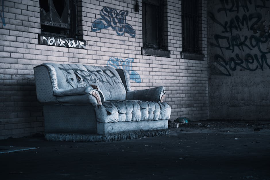 A worn, light blue fabric-upholstered sofa with visible tears and patches of missing material, positioned on a dark concrete floor inside an industrial-like space with exposed brick walls that are covered in blue and black graffiti. The sofa's armrests show signs of damage, and its cushions appear flattened and aged. Behind the sofa, there are three small, barred windows with black security bars, and a clutter of objects including cardboard boxes and plastic wrapping materials scattered on the floor nearby. The environment is dimly lit, with shadows cast across the scene, suggesting a setting involved in furniture disposal or a furniture collection process as part of a house removal or relocation service, as provided by Man with Van West Drayton.