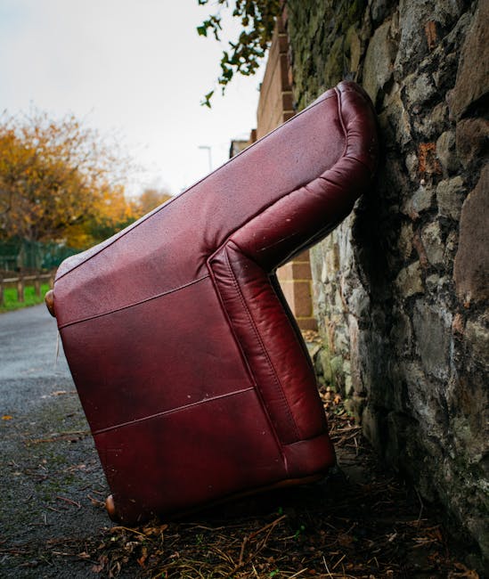 A red leather armchair has been placed on the ground outside, leaning against a stone wall in an outdoor setting. The chair is positioned with the backrest facing the wall, with its armrests and cushioned seat visible. It is situated on a paved surface, with some scattered leaves and debris nearby, indicative of a street or pathway in a residential area. In the background, a portion of a brick building and a green fence are visible, along with trees displaying autumn foliage. The lighting appears natural, suggesting daytime. The image is related to house removals or furniture transport, with the chair being prepared for loading or removal, demonstrating the kind of furniture handling involved in home relocation services provided by Man with Van West Drayton.