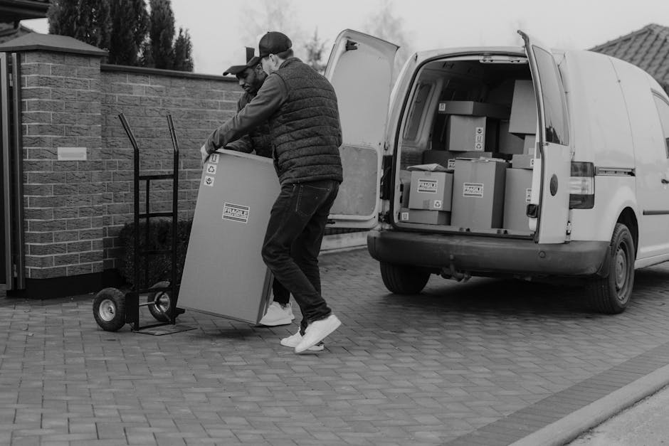 Two men are engaged in a home relocation process outdoors in West Drayton. One is lifting a medium-sized cardboard box, which has protective packaging and label markings, while the other pushes a small hand truck with another box on it. They are positioned on a paved driveway next to a brick wall and a white van with its rear doors open, revealing more packed cardboard boxes and possibly household items inside. The men wear casual clothing, including jackets, jeans, and caps, and appear focused on loading the boxes onto the vehicle as part of a furniture transport or packing and moving operation. Equipment such as trolleys and packaging materials are visible, emphasizing an organized moving process, with the setting taking place during daylight hours, capturing a typical van-loading scene associated with professional removals services, such as those offered by Man with Van West Drayton.