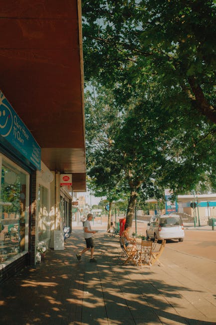 A street scene showing the entrance to a shop with a blue signboard, part of which reads 'Post Office', under a brown canopy. The sidewalk is shaded by a large leafy tree whose branches extend over the pavement. Two people are present: one person, dressed in a white shirt and dark shorts, is walking along the sidewalk, while another person, nearer to the shop, appears to be carrying or moving items, possibly related to a home relocation. Several cardboard boxes and packing materials are placed near the shop entrance, indicating packing or moving activities. A small table with chairs is situated outside, under the shade of the tree, suggesting a casual outdoor seating area. In the background, parked vehicles and a few more shops are visible, with clear daylight illuminating the scene. The environment reflects a typical busy high street, relevant to house removals and furniture transport services provided by Man with Van West Drayton.
