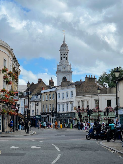 The image shows a street scene in West Drayton with two adjacent buildings featuring traditional timber-framed facades, painted in a combination of black and white, and brown and beige, respectively. The ground floors of these buildings include a café or restaurant with large display windows, and outdoor seating with tables and chairs where two elderly people are seated, engaged in conversation. A large black lamppost stands in the pavement near the seating area. In the foreground, part of a road with a marked cycle lane is visible, with a pedestrian walking past on the pavement. Bright, natural daylight illuminates the scene, with green tree leaves hanging over the top of the image, partially shielding the view. The overall setting suggests a busy street likely involved in home relocation activities such as furniture transport or packing during a moving process, supported by Man with Van West Drayton’s local presence in removals and home moving services.
