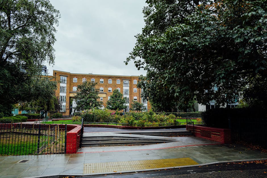A street-level view of an apartment building in West Drayton with a paved pedestrian area leading to a staircase that provides access to a residential stairwell. The staircase is made of concrete and has black metal railings on both sides, with some red brick planters at the base. To the left and right of the stairs, there are green lawns and mature trees providing natural shade and privacy, with dense foliage on the right. The background features a multi-storey brick residential building with white-framed windows, and the scene appears to be overcast with diffused natural light. The location suggests it could be an ideal setting for house removal or furniture transport services, such as those offered by Man with Van West Drayton, especially in terms of stair access considerations for home relocations or apartment moves.