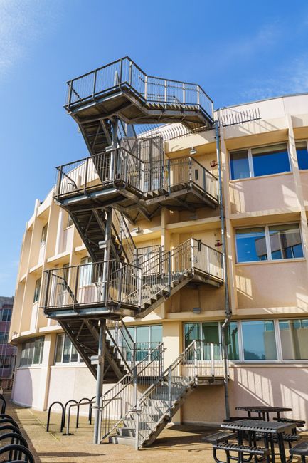 An outdoor metal staircase with four landings and black safety railings is attached to a multi-storey residential building with cream-colored exterior walls and large windows. The staircase provides access to different floors via exterior walkways. The sky is clear and blue, and the area at the base of the staircase features a paved courtyard with black bicycle racks, a picnic table, and a bench. This setting illustrates typical access points used during home relocation or furniture transport in a flat residential setting, where exterior staircases often facilitate movement of packed boxes, furniture, and appliances during a removal service. Man with Van West Drayton specialises in house removals and can assist with logistics related to upstairs and downstairs access, ensuring secure handling and transport of belongings during the loading process, especially for flats with external stair access.