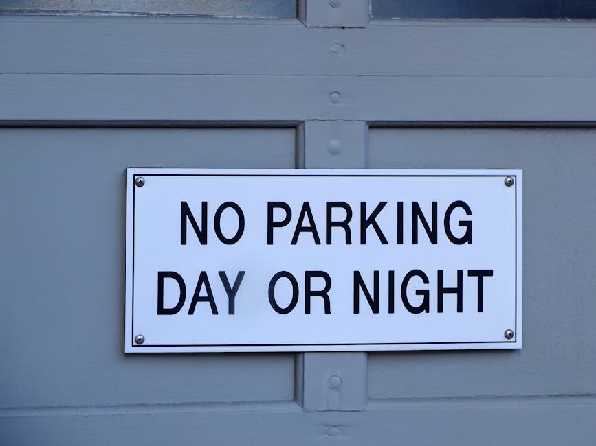 A rectangular white metal sign with black uppercase text reading 'NO PARKING DAY OR NIGHT' is mounted on a light grey wooden exterior wall, likely part of a residential or commercial building. The sign is secured with four screws, one in each corner. Behind the sign, the wooden paneling features horizontal grooves and visible fasteners, indicating a sturdy construction. The sign's placement suggests a designated area where parking is prohibited at all times to facilitate house relocation or furniture transport processes, consistent with typical removals and moving services in West Drayton. The lighting appears natural, illuminating the sign clearly for visibility during daytime use, supporting effective parking regulation and smooth logistics for home removals operated by Man with Van West Drayton.